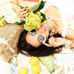 Woman lying on a blanket with white sunglasses, surrounded by flowers, a basket, and lemons, enjoying a picnic.