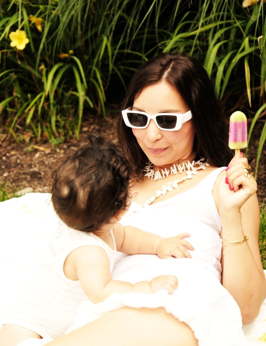 Woman in white sunglasses enjoying a colorful popsicle while playfully interacting with a baby outdoors.