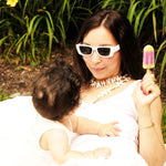 Woman in white sunglasses enjoying a colorful popsicle while playfully interacting with a baby outdoors.