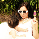 Woman in white sunglasses enjoying a colorful popsicle while playfully interacting with a baby outdoors.