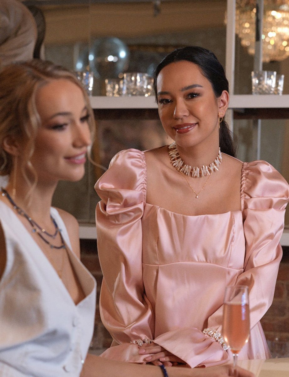 Two women wearing elegant outfits and jewelry, enjoying a social gathering with drinks.