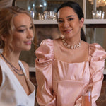 Two women wearing elegant outfits and jewelry, enjoying a social gathering with drinks.