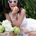 Woman in white outfit and sunglasses, smiling with a lemon, surrounded by flowers and a picnic basket.