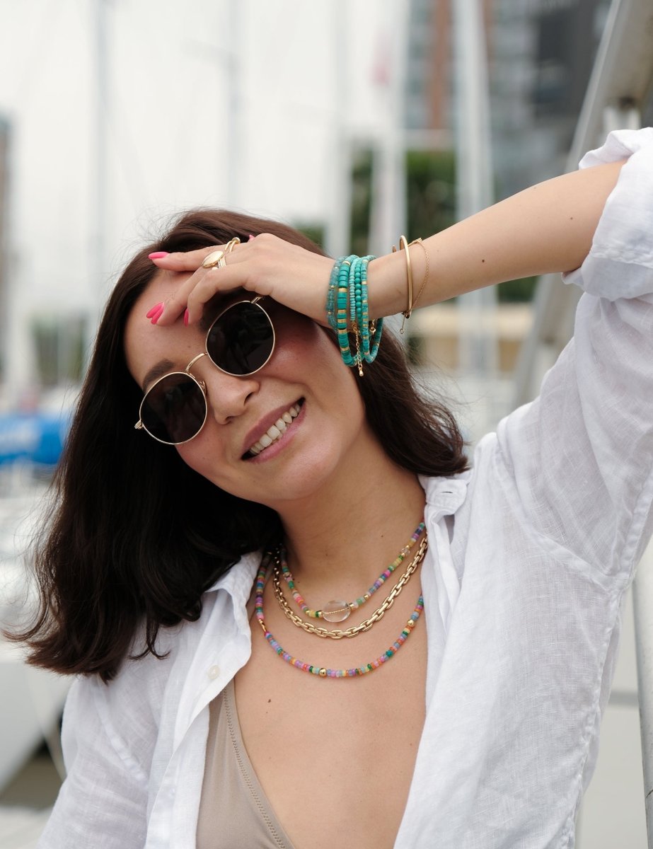 Model wearing stylish sunglasses and layered necklaces in a marina setting, showcasing vibrant colors and accessories.