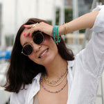 Model wearing stylish sunglasses and layered necklaces in a marina setting, showcasing vibrant colors and accessories.