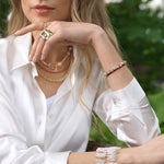 Model wearing layered jewelry including necklaces and bracelets, showcasing elegant accessories against a natural background.
