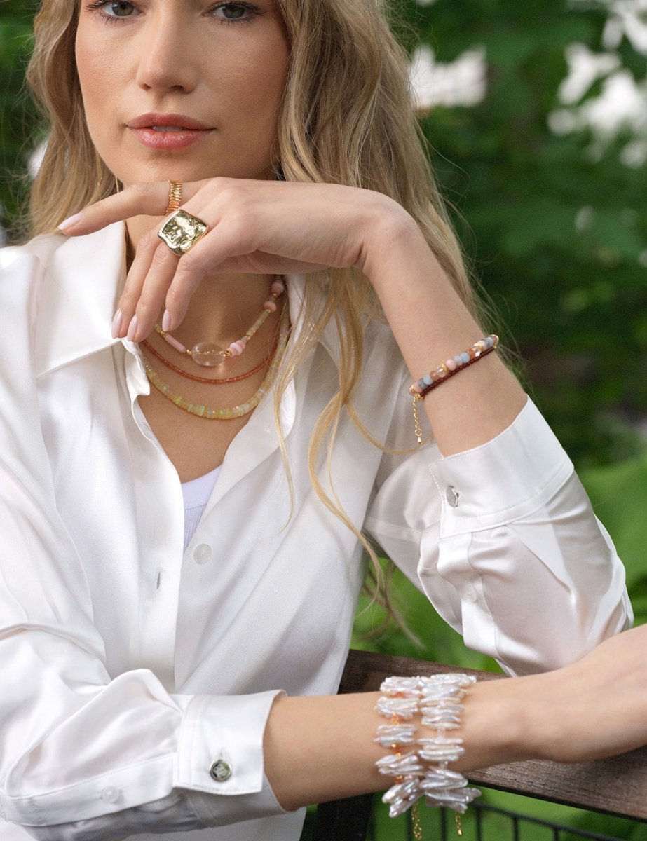 Model wearing layered jewelry including necklaces and bracelets, showcasing elegant accessories against a natural background.
