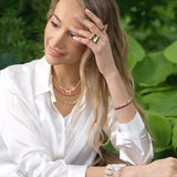 Woman wearing layered jewelry and a ring, showcasing elegance and style in a natural setting.